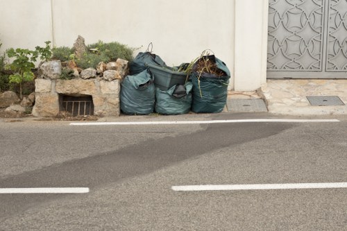 A row of three large, dark green refuse collection bags filled with mixed waste is placed against a white exterior wall on the edge of a paved street in Hayes, with a small garden area containing rocks and greenery on the left, and a decorative gray metal gate on the right. The bags appear to be made of durable plastic, with some loose material and plant debris visible at the openings. The street surface is smooth with painted white lines marking parking bays, and a small metal drain cover is embedded near the wall. The overall setting suggests a typical rubbish disposal scene in a residential or commercial area in Hayes, where Commercial Waste Removal Hayes offers rubbish collection and disposal services, with the bags positioned on the curbside, ready for collection.