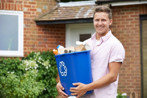 A young man with short light brown hair and wearing a light pink short-sleeved shirt is standing outdoors in front of a brick house with a white-framed window and a green garden. He is smiling and holding a blue recycling bin made of plastic, filled with various types of waste including cardboard boxes, paper, and plastic bottles. The bin features the internationally recognized recycling symbol in white on its side. The setting appears to be a residential area, with greenery and bushes visible in the background, suggesting a typical suburban environment. The scene is well-lit with natural daylight, emphasizing the clean and orderly appearance of the surroundings. This image is relevant for a rubbish removal service such as Commercial Waste Removal Hayes, serving local clients in the Hayes area, UK, focusing on sustainable waste collection and disposal practices.