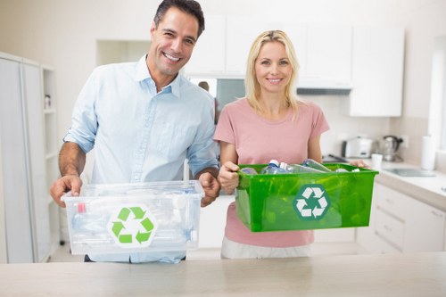 A middle-aged man and woman stand side by side in a bright, modern kitchen, each holding a reusable recycling bin. The man, dressed in a light blue shirt, holds a clear plastic bin with a green recycling symbol, revealing a few crumpled paper and plastic bottles inside. The woman, wearing a pink T-shirt, carries a sturdy green bin also marked with a recycling symbol, containing empty plastic bottles and paper packaging. Both are smiling and facing forward, positioned in front of a white countertop with integrated appliances, and a clean, minimalist kitchen environment with white cabinets and a grey backsplash in the background. The scene emphasizes sustainability, recycling practices, and environmentally conscious waste management at home, subtly supporting local waste removal services in Hayes and surrounding areas. The overall lighting is natural, enhancing the clarity of the scene, with the focus on their engagement in eco-friendly waste separation activities.
