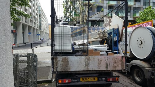 A small flatbed utility truck parked on a city street corner, loaded with a large pile of mixed rubbish and waste materials. The waste includes white and light grey panels, possibly broken furniture or construction debris, stacked haphazardly on the truck's flatbed. The truck’s metal sides show signs of rust and wear, with some areas covered in dirt. Behind the load, a black metal mesh partition divides the cargo area from the driver's cab. To the right, a white cycle wheel, possibly part of equipment or a small container, is visible. The background features a modern residential or commercial building with white facades and large windows, and a traffic light at the intersection. The scene is set during daylight hours with overcast weather, typical of urban rubbish removal operations, and suggests that Commercial Waste Removal Hayes offers local waste collection services in the Hayes area, which is located in West London.